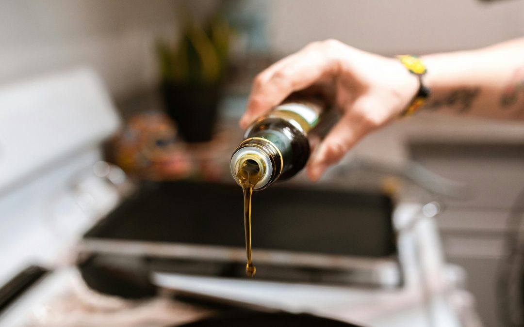 Hand holds oil bottle over pan on white stove while cooking oil begins to pour out.